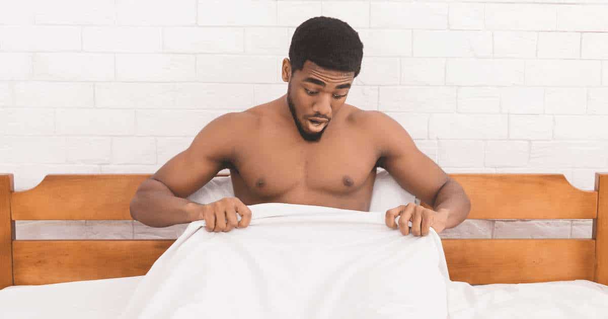 A man on a bed expressing astonishment at his intimate area concealed by the blanket.